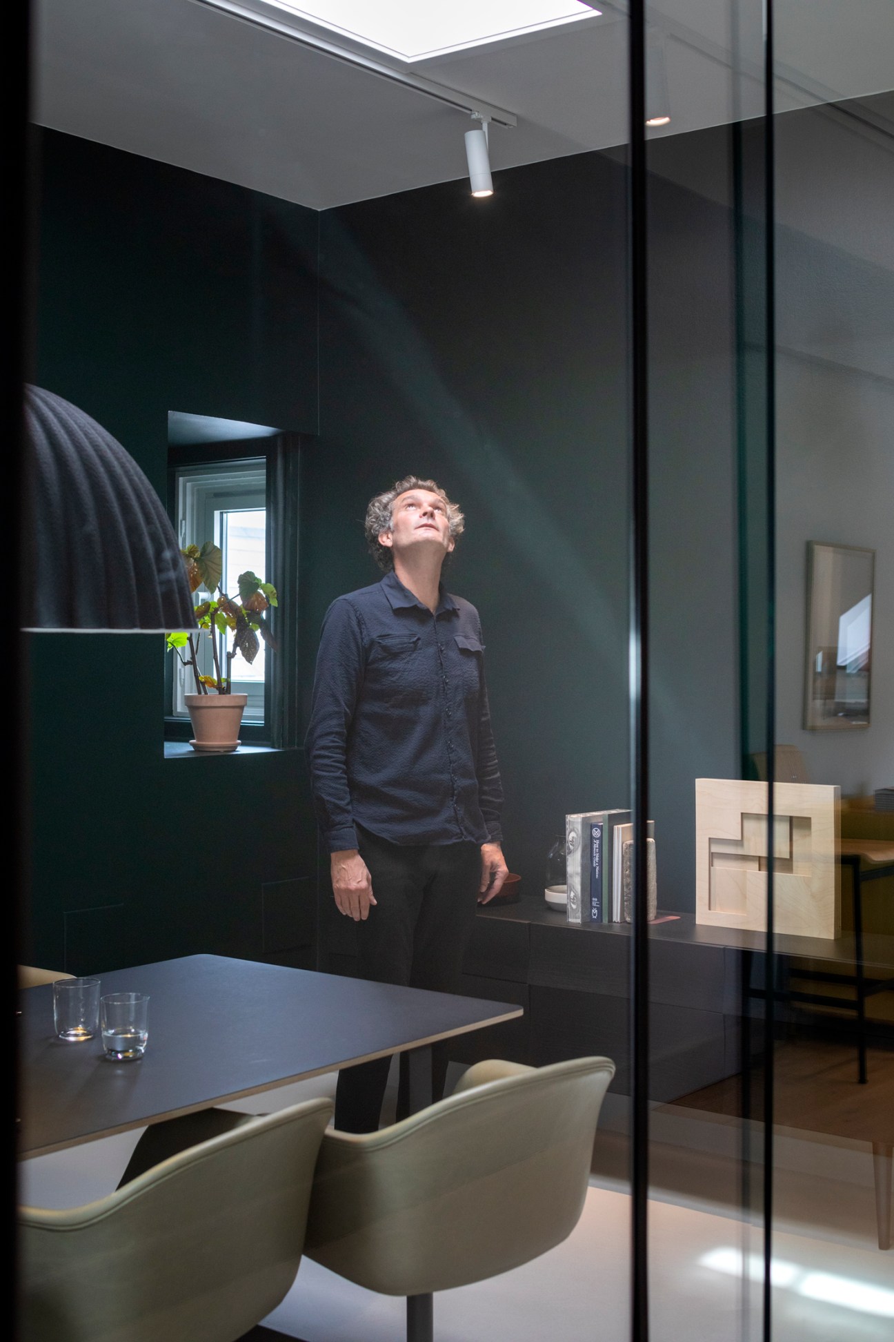 Man in navy shirt looking up at skylight in modern dark-walled office space with dining table and chairs