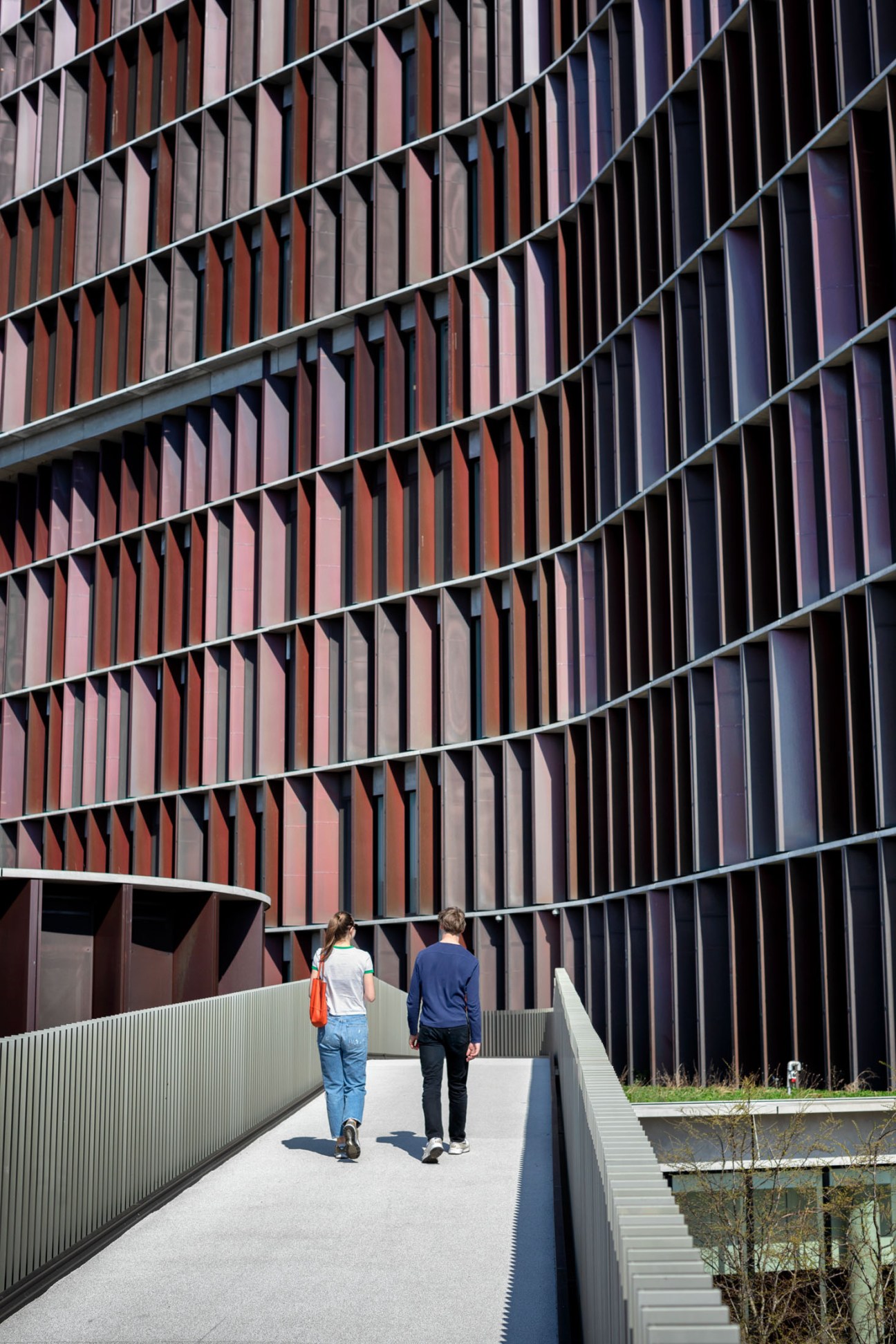 Two pedestrians walking on an elevated concrete pathway past a building with a weathered steel undulating facade.