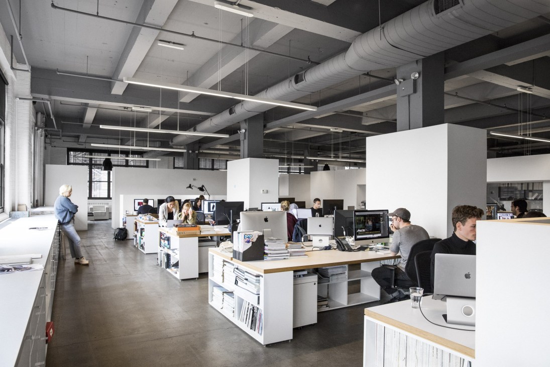 Open-plan creative office with white storage units, wood desks, and employees working on computers under industrial ceiling.