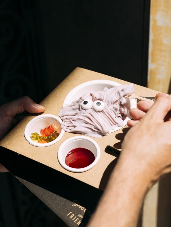 Person holding a wooden board with whimsical shaved ice cream creature with candy eyes and condiment bowls.