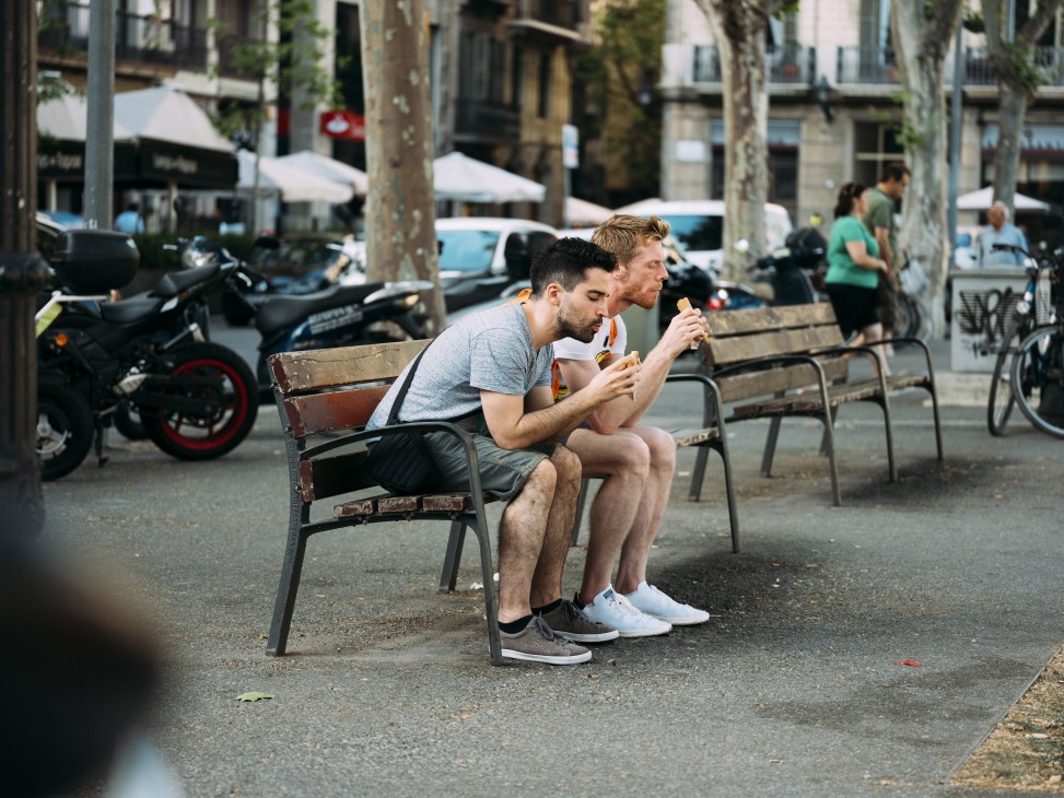 Two men sitting on chairs eating ice cream on a Barcelona street lined with trees and scooters.
