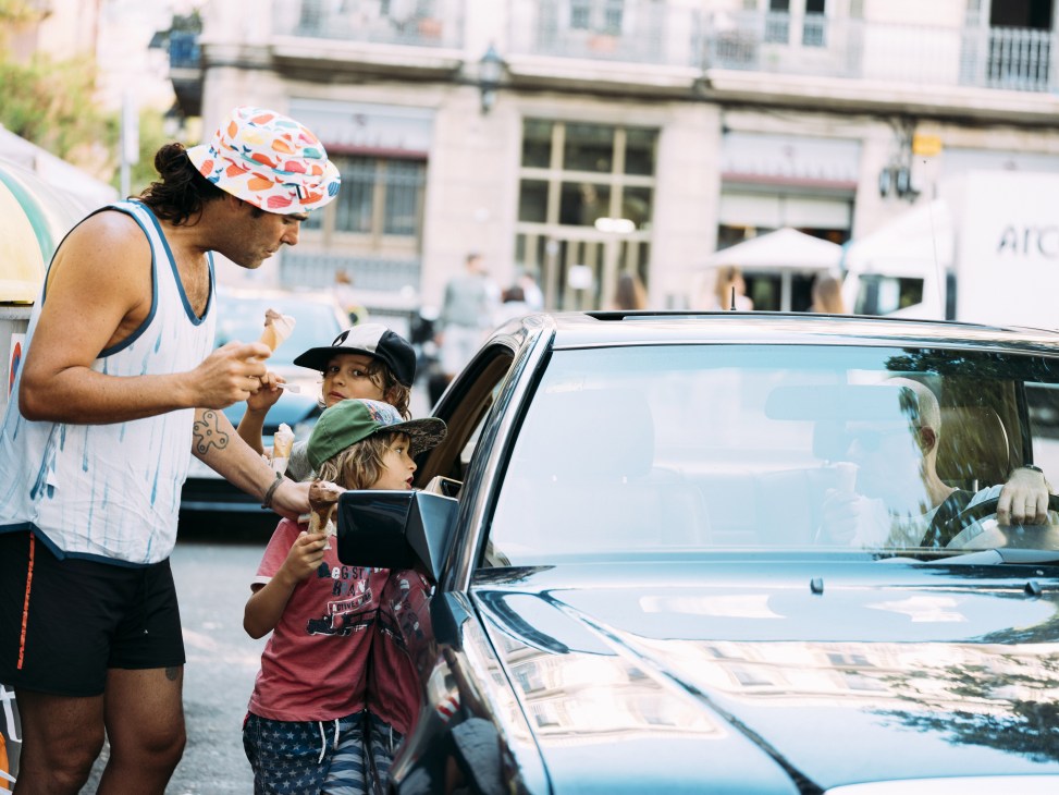 Man in colorful cap serving ice cream to two children at car window on city street.