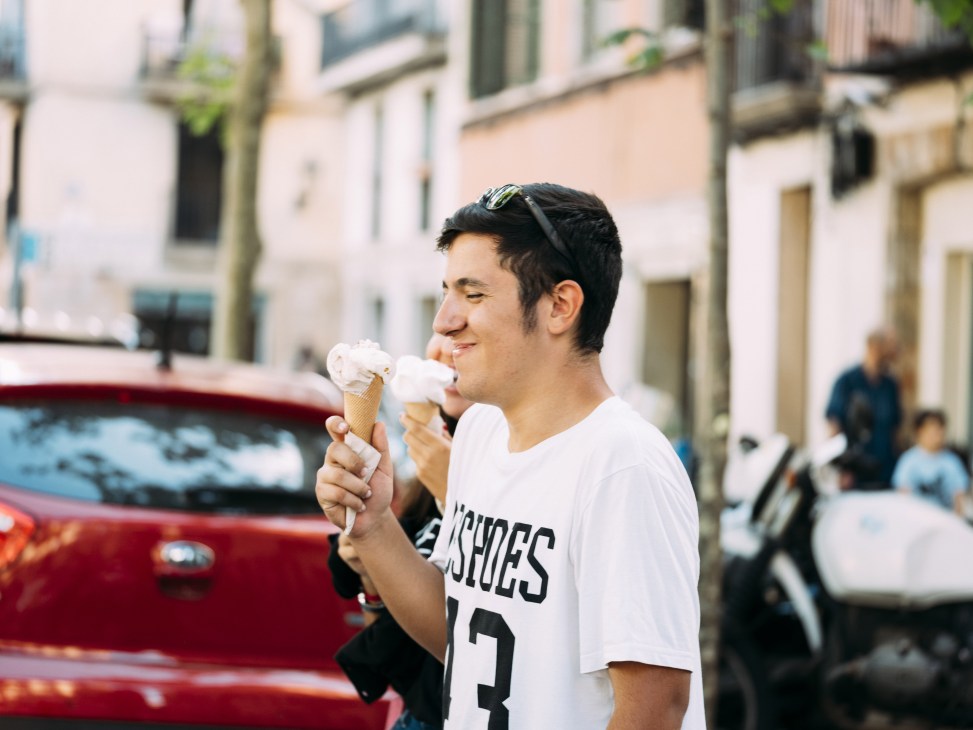 Young man in white t-shirt eating ice cream cone on a sunny urban street.