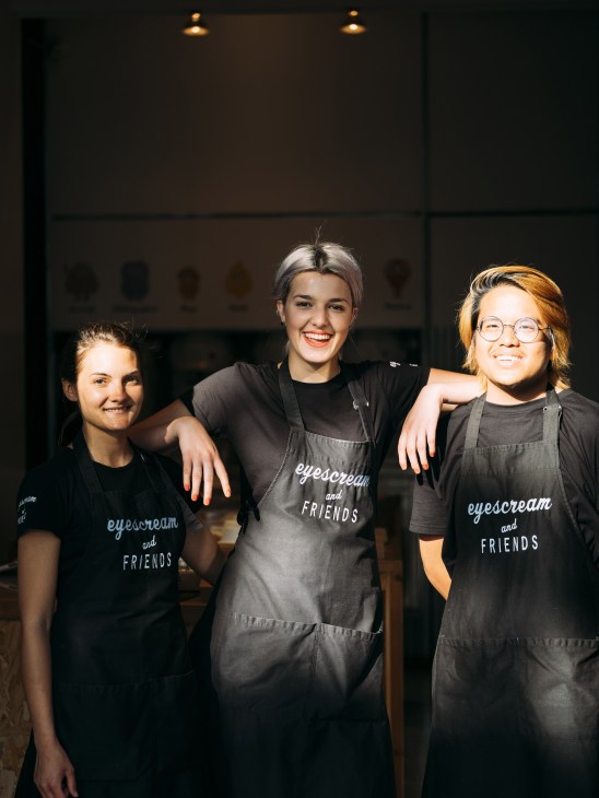 Three staff members wearing 'eyescream and Friends' aprons smile at the camera in an ice cream parlor.