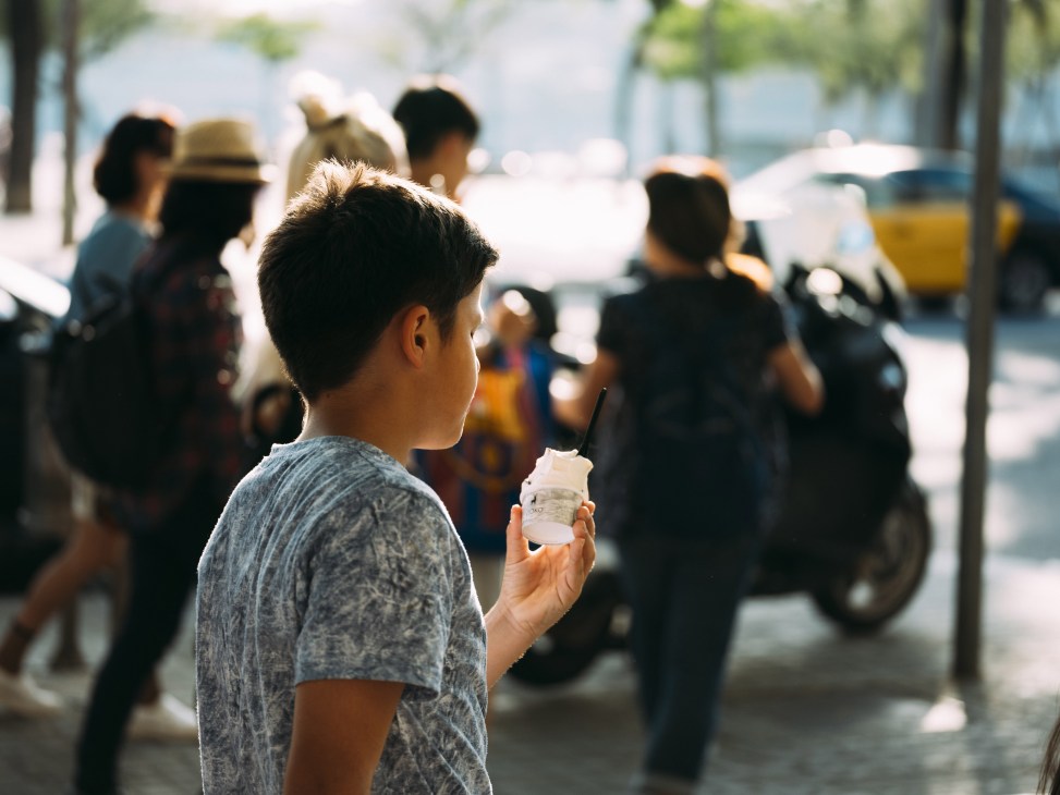 A young boy in a light blue shirt holds a small ice cream or frozen treat while standing on a busy urban street with blurred pedestrians and vehicles in the background.