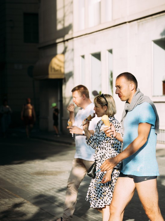 A man in a blue striped shirt and shorts holds a young girl eating ice cream while walking past a white building.