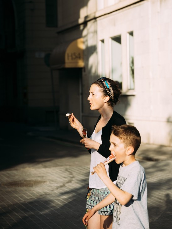 Woman and young boy eating ice cream cones on a sunny street in front of white buildings.