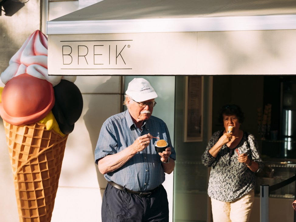 An elderly man in a white cap and blue shirt eats ice cream outside a Breik ice cream shop while a woman enjoys gelato nearby.