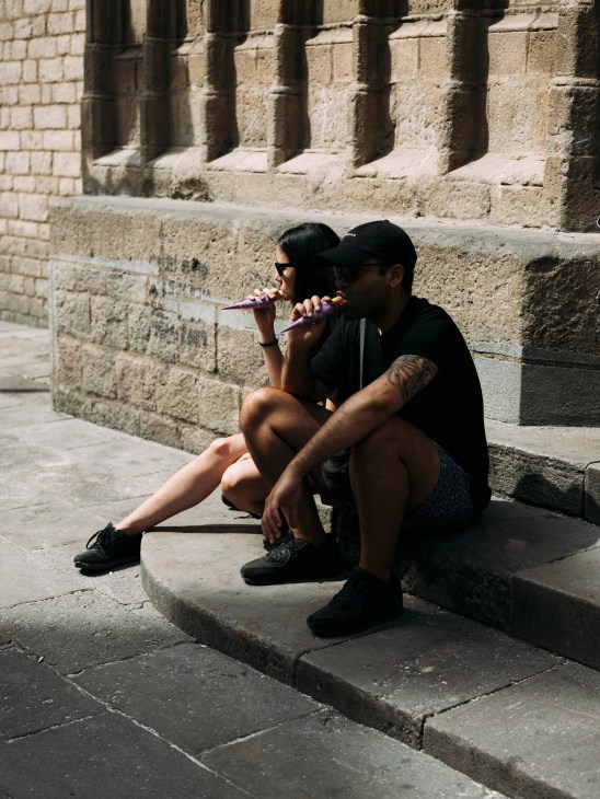 Two people sitting against a stone wall eating ice cream from cones in an urban setting.
