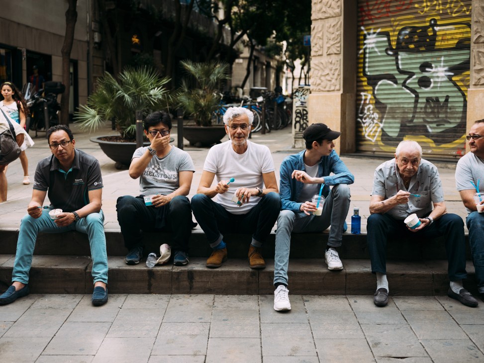 Group of men of various ages sitting on a step enjoying ice cream in a sun-lit Barcelona street.