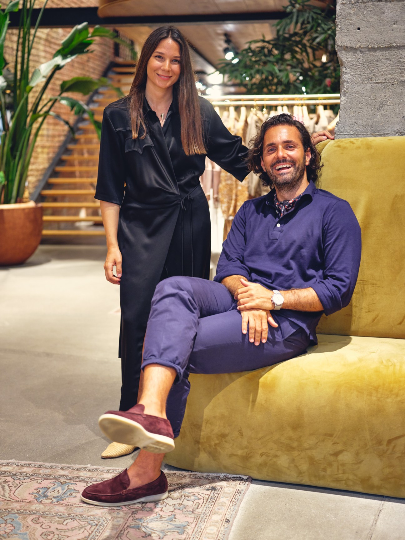 Sandra Sandor and Peter Baldaszti pose together in Nanushka's Budapest shop on a mustard-yellow couch.
