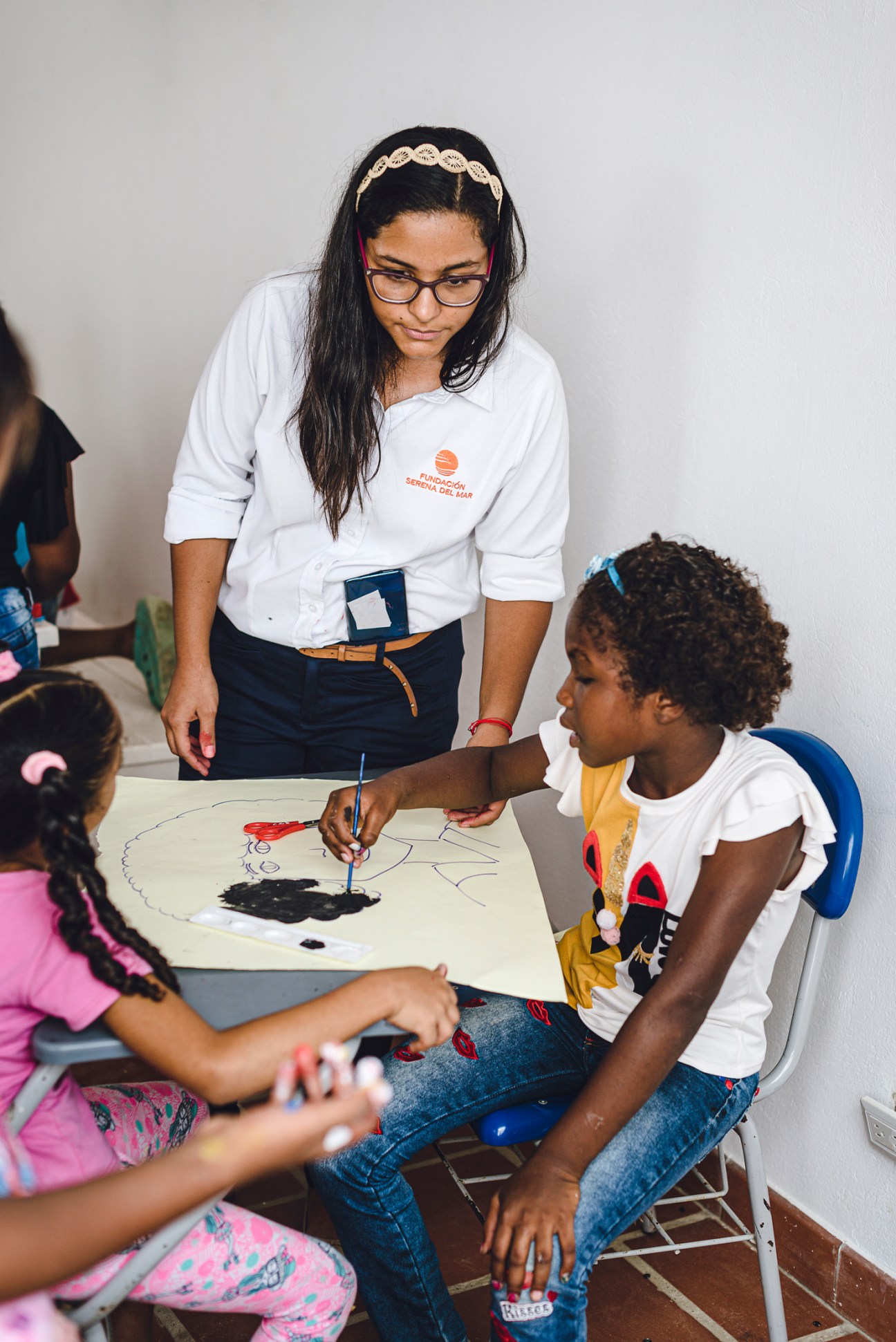 Woman in white shirt helping children with art project at Fundación Serena del Mar classroom