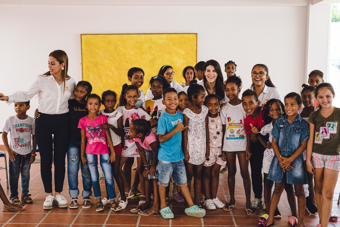 Group of smiling children and adults posing together in a classroom with white walls and yellow artwork