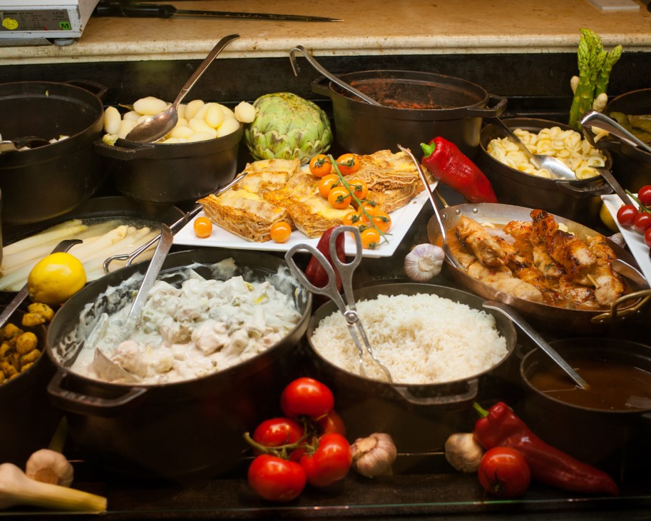 A deli counter display featuring cooked foods in black pans including roasted meat, artichoke, potatoes, rice, and fresh vegetables with serving utensils.