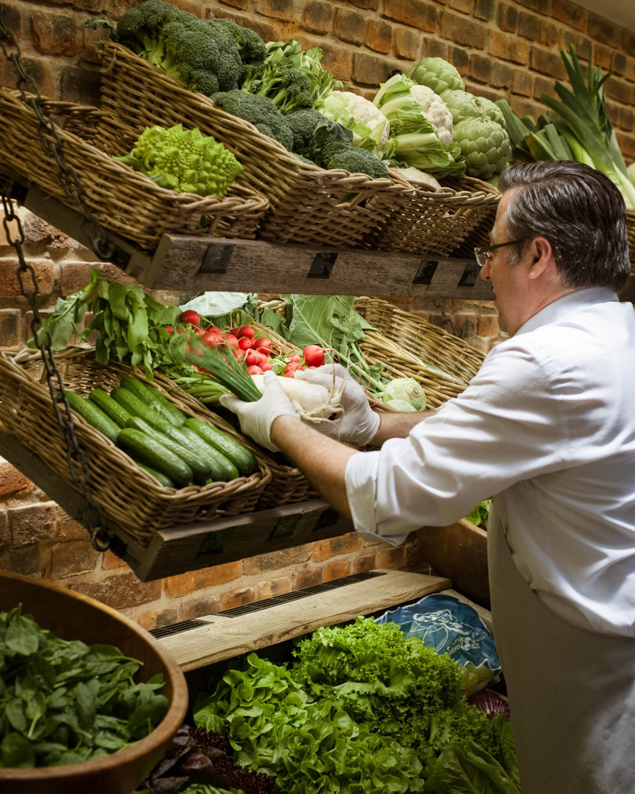 A man in white gloves and shirt arranges fresh produce in wicker baskets against a brick wall at Käfer supermarket.