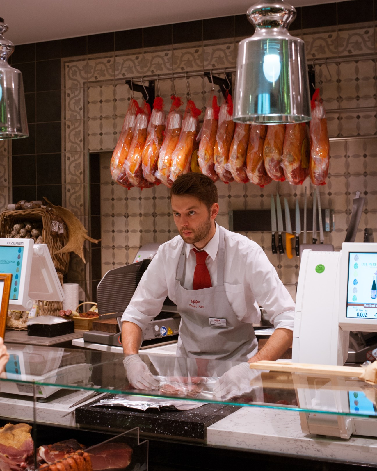 A deli counter assistant in white uniform and red tie stands behind a glass counter at Käfer supermarket in Munich.