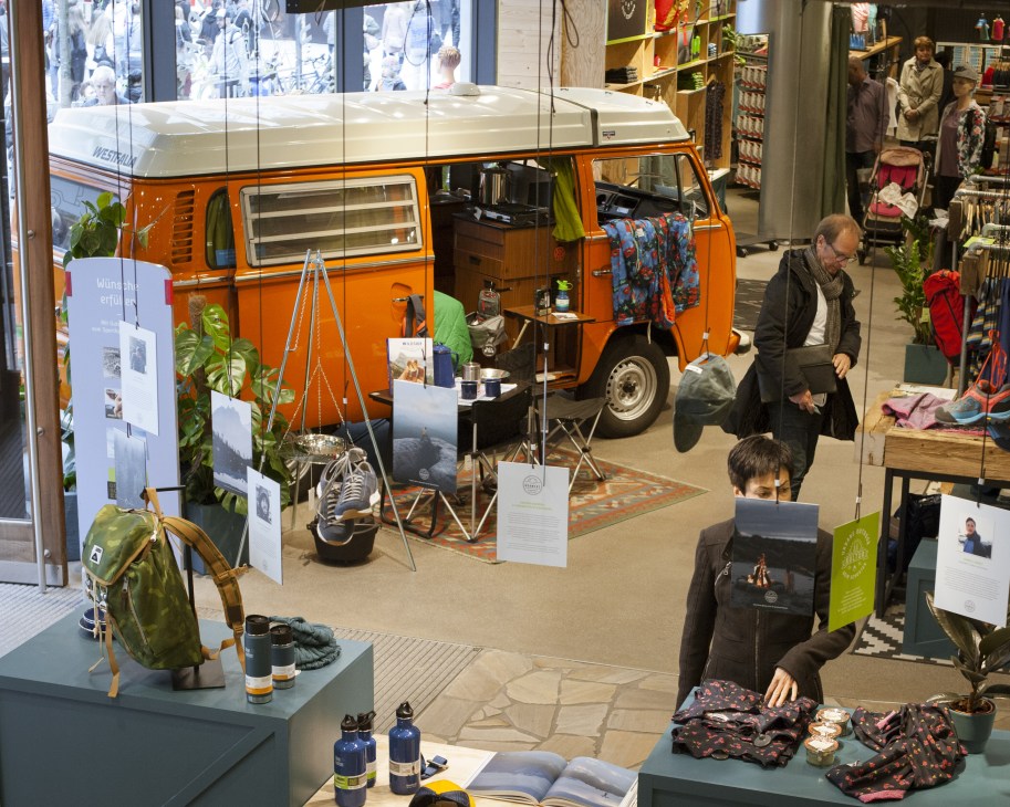 Orange vintage camper van displayed inside Sporthaus Schuster sporting goods shop with camping equipment and customers browsing.