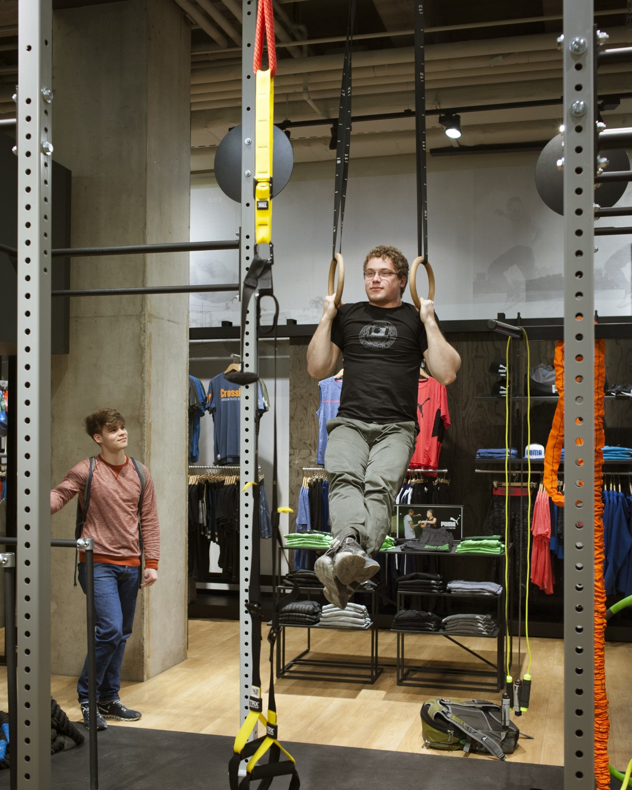 Man performing pull-ups on gymnastics rings in Sporthaus Schuster's workout testing area while a child watches.