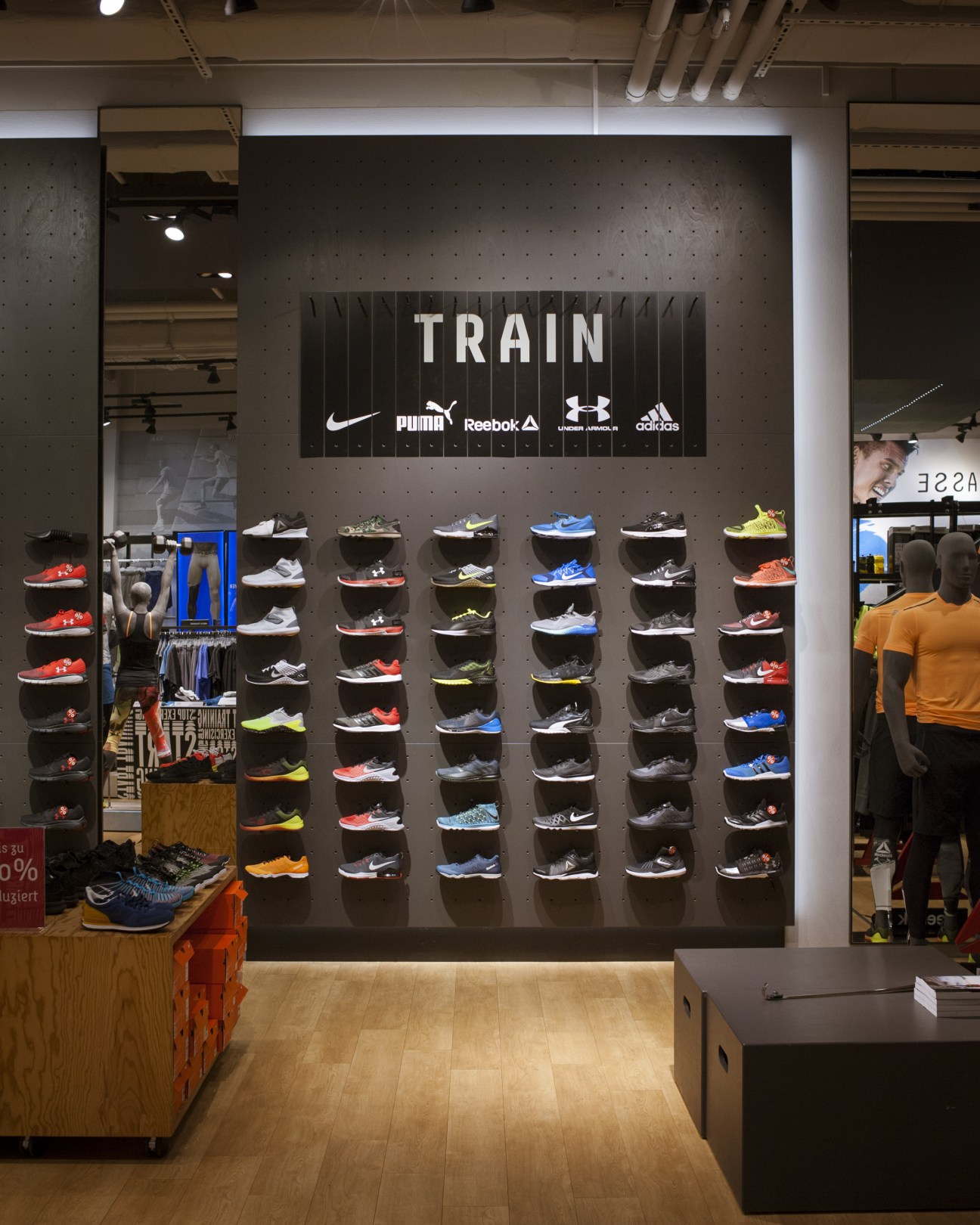 Sporthaus Schuster storefront display wall with "TRAIN" signage and athletic shoes arranged on shelves featuring Nike, Puma, Reebok, Under Armour and Adidas branding.