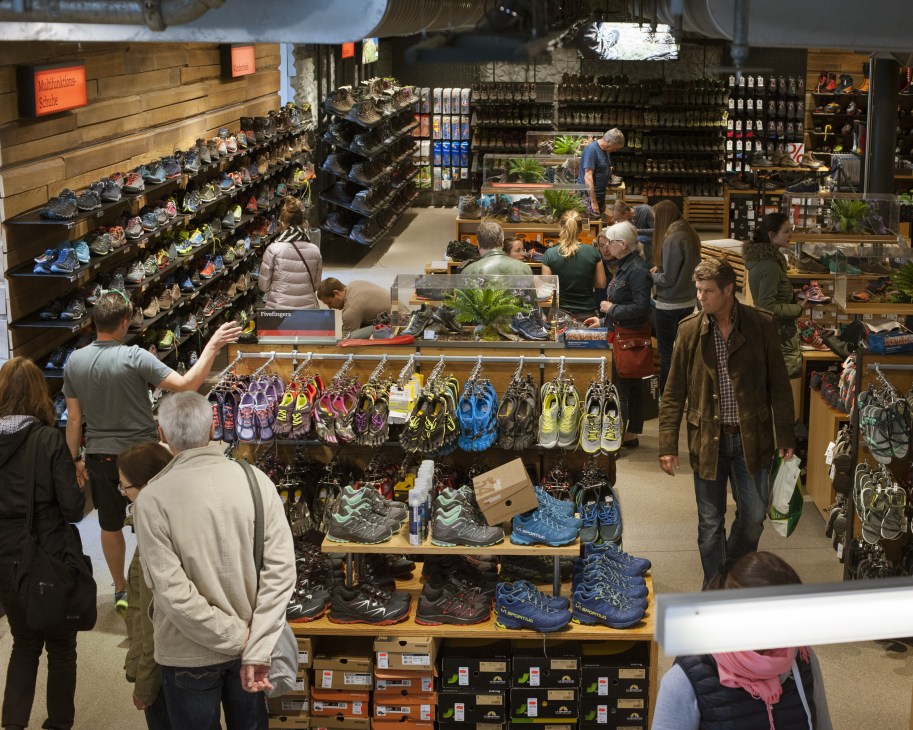 Interior view of Sporthaus Schuster showing customers browsing hiking boots and outdoor gear across multiple display tables and shelves.
