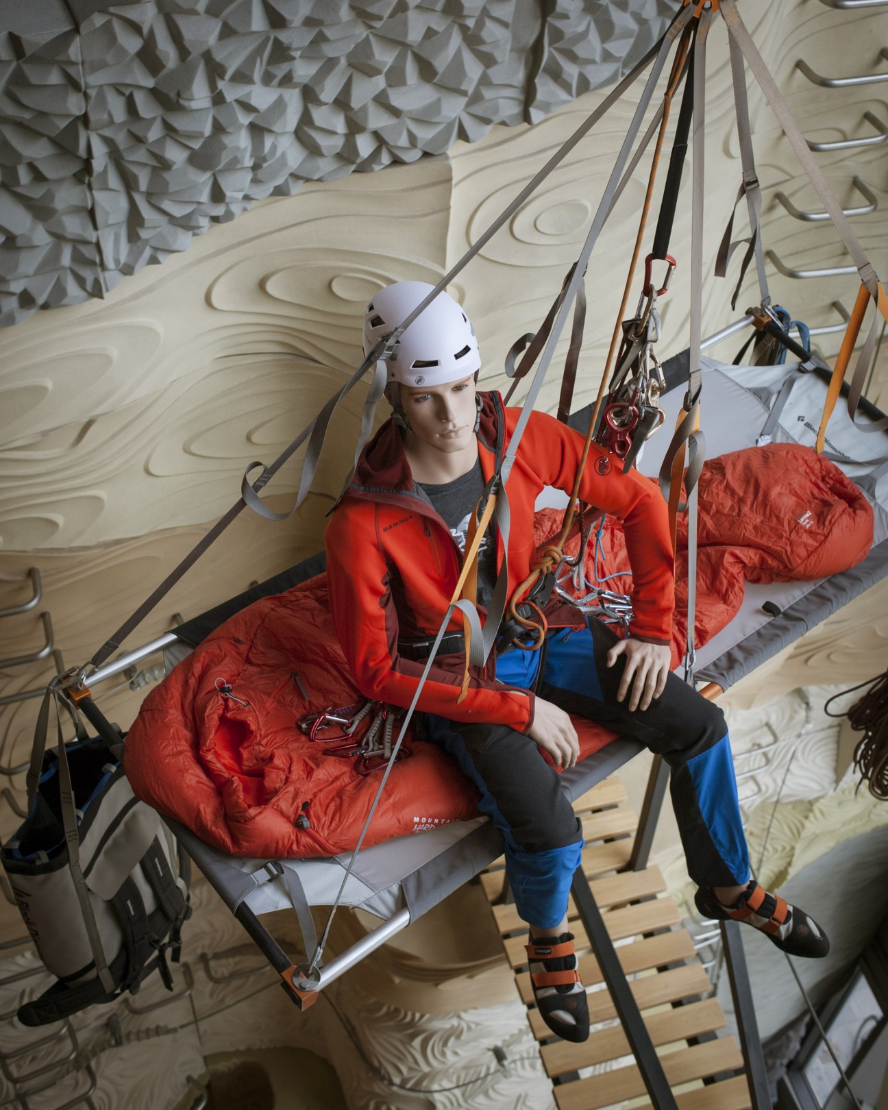 Mannequin in red climbing gear and white helmet suspended on ropes against textured climbing wall.