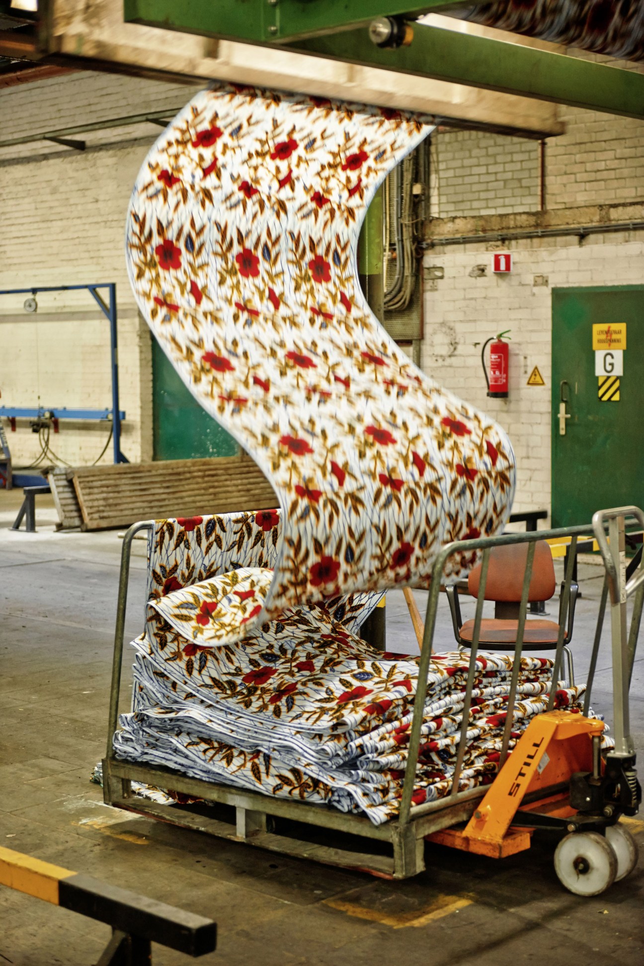 Rolled Vlisco wax-printed cotton fabric with red flowers and gold leaf pattern displayed in a factory warehouse.