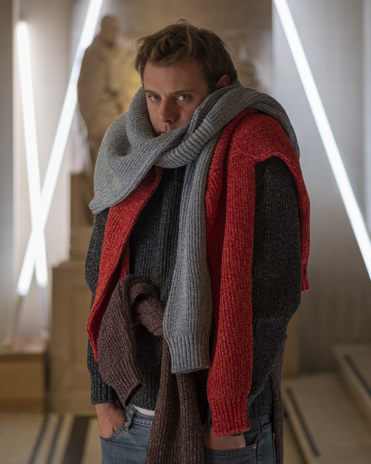 Man wearing layered knit sweaters in grey, red and black with a grey scarf in a modern corridor with neon lighting.