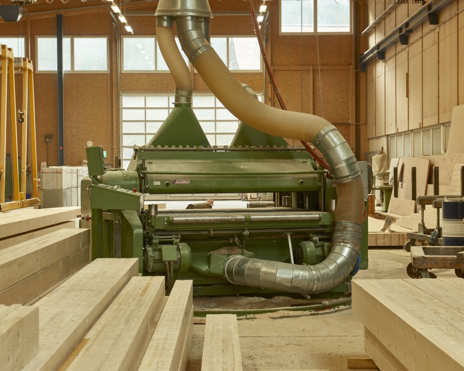 Industrial planing machine with green metal frame and flexible dust collection ducts in a wooden workshop.