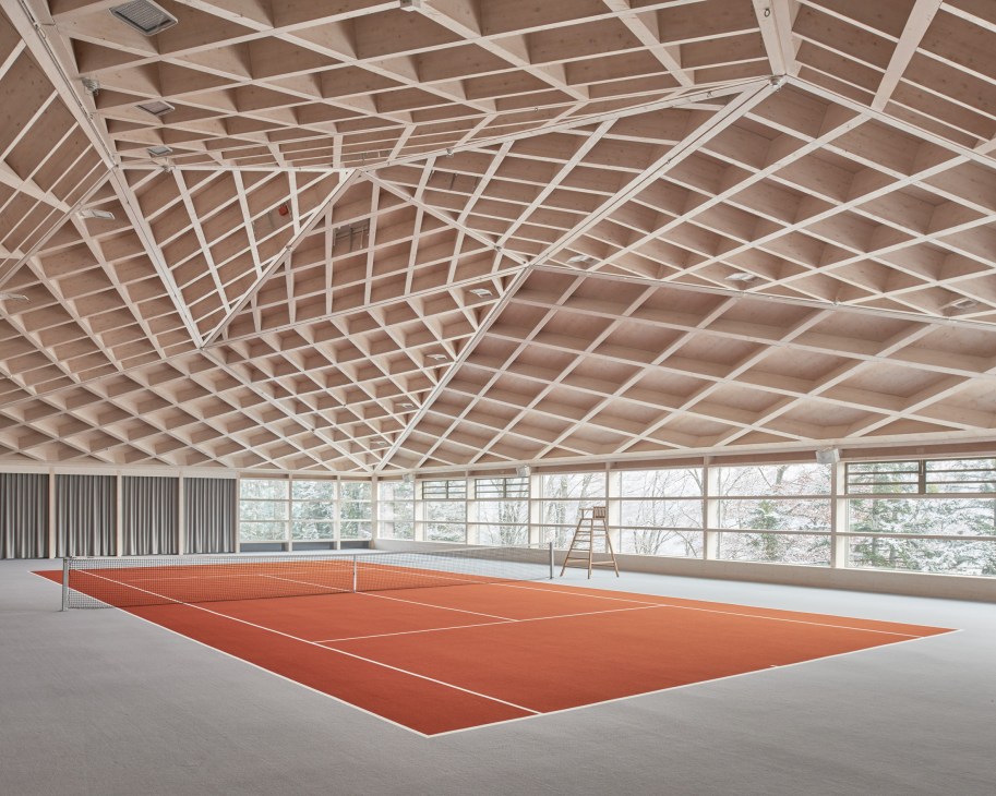 Modern indoor tennis court with orange clay surface, angular white timber ceiling, and floor-to-ceiling windows overlooking mountains.