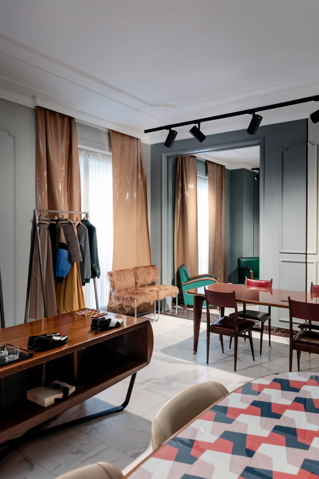 Modern showroom interior with mid-century furniture, tan curtains, and turquoise chair displaying designer clothing on racks.