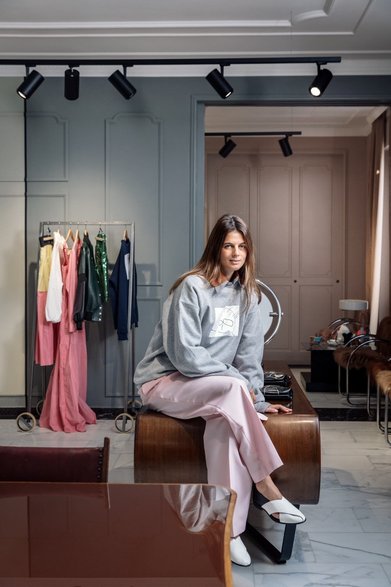 Woman sitting in a modern showroom wearing gray sweatshirt and pink trousers, with colorful clothing on a rack behind her.