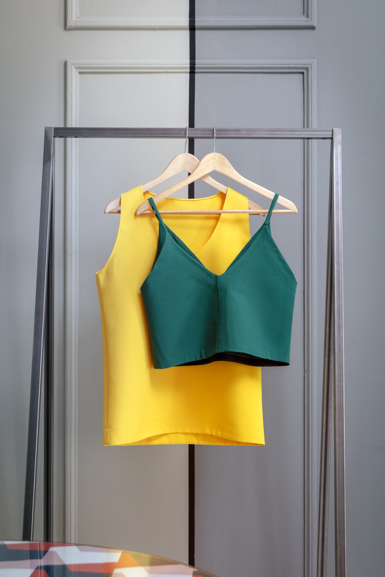 Yellow and teal sleeveless tops displayed on wooden hangers against a paneled wall.