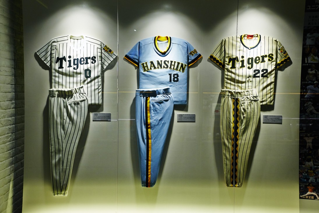 Three Hanshin Tigers baseball uniforms displayed in a museum case, including pinstriped home jerseys and a blue Hanshin away uniform.