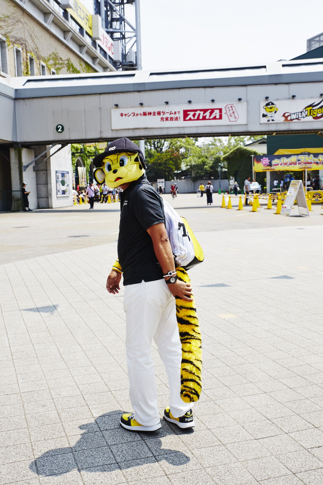 A fan in full Hanshin Tigers costume with yellow tiger mascot head and striped uniform poses outside the stadium.