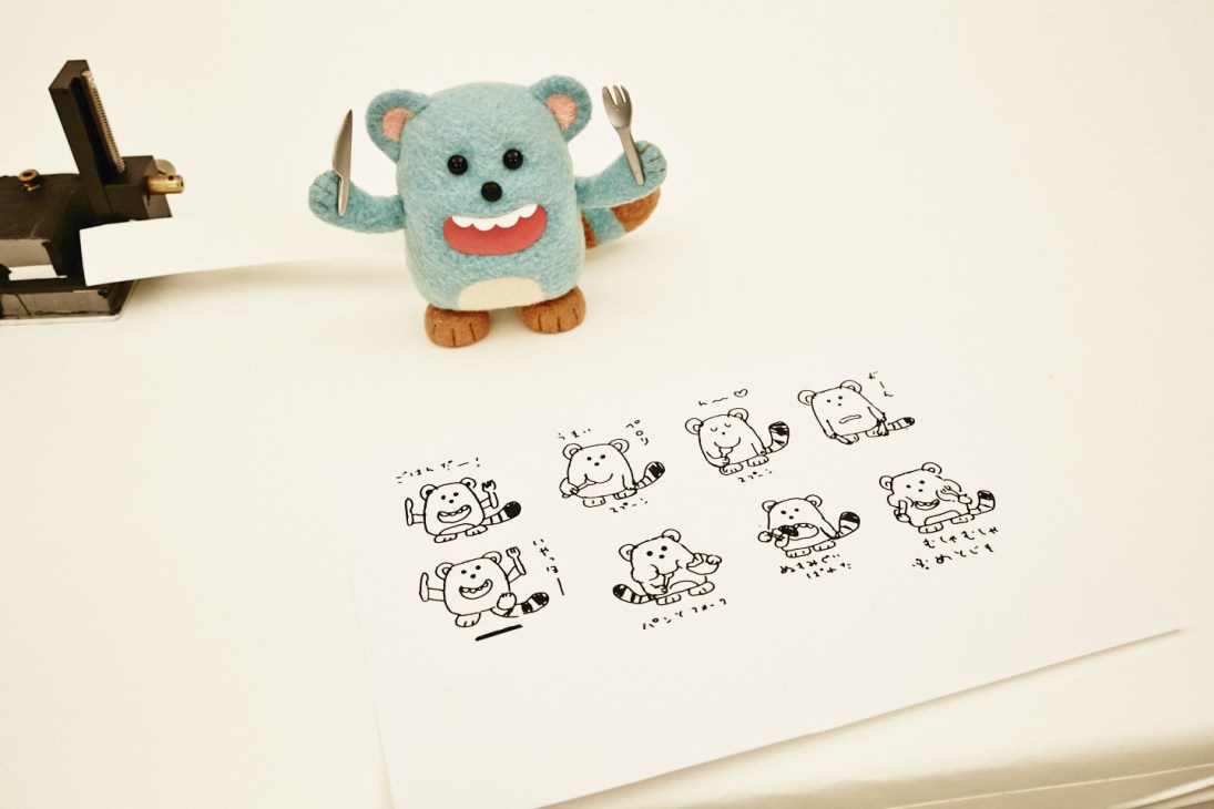 A turquoise felt puppet with red mouth and utensils positioned above hand-drawn character sketches on paper.