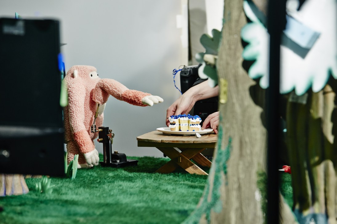 Stop-motion animator adjusting a pink puppet character on a miniature set with tiny cake props and artificial grass.