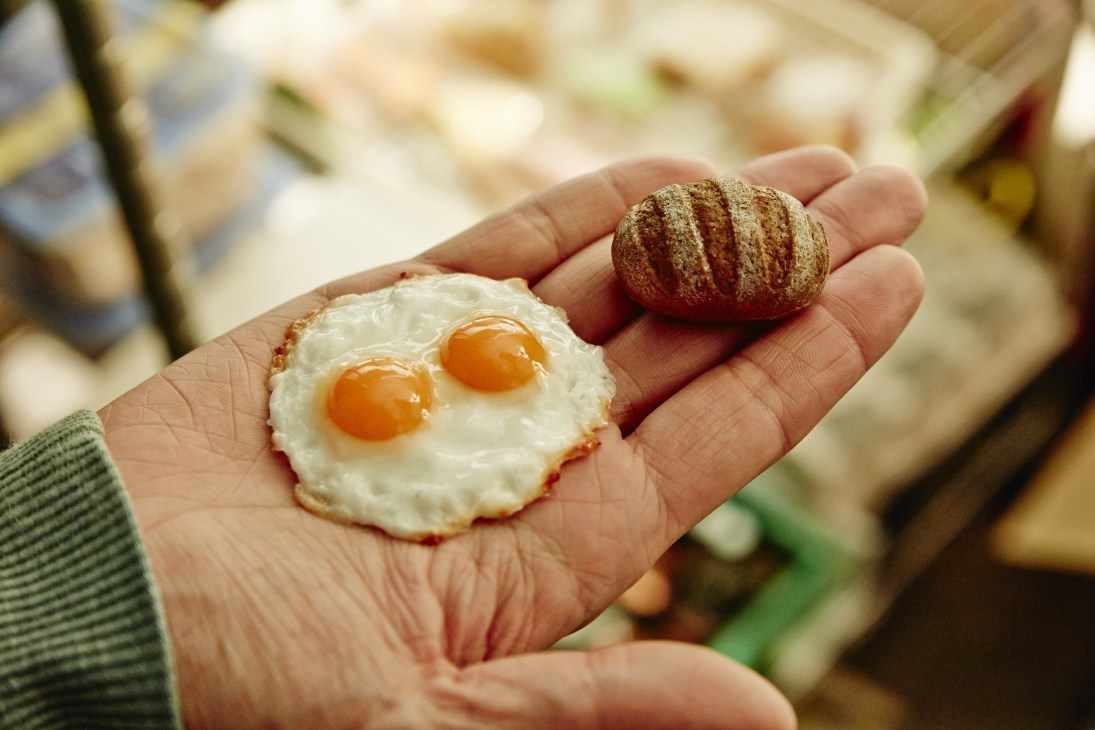 Hand holding miniature plastic food props including sunny-side up eggs and a bread roll for stop-motion animation.