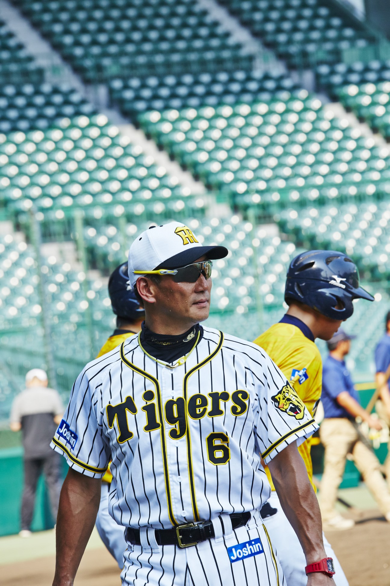 Hanshin Tigers shortstop Fumiya Hojo in pinstriped uniform and cap standing in the dugout with bat, stadium seats blurred behind.