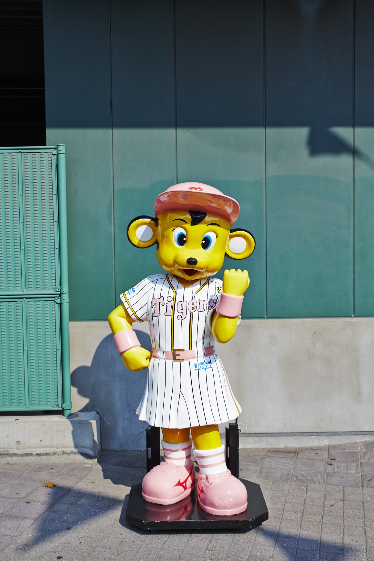 Hanshin Tigers mascot To-Lucky in pinstriped uniform and red cap with pink shoes, standing against a teal-paneled stadium backdrop.