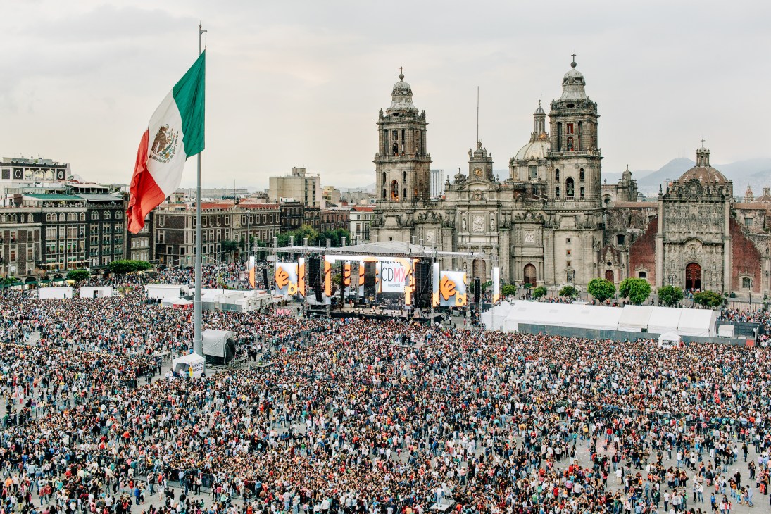 Crowd of thousands gathering in Mexico City's Zócalo main square with stage, Mexican flag, and colonial cathedral in background.