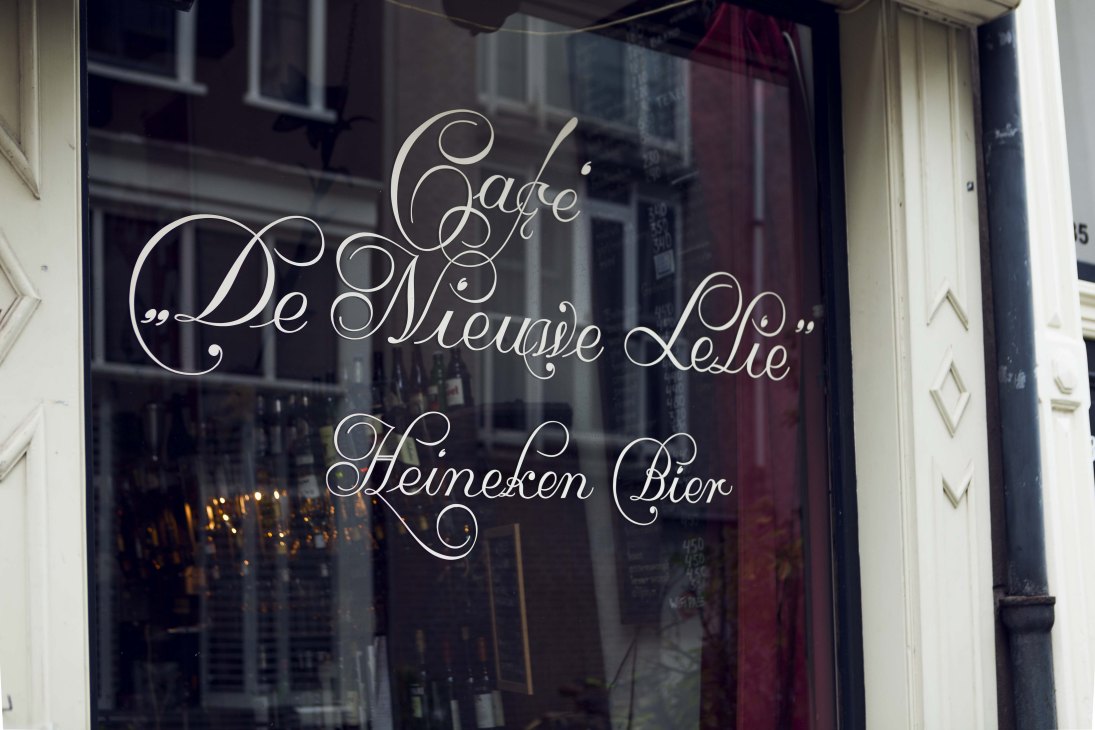 Hand-painted Amsterdamse Krulletter script on café window reading 'Café de Nieuwe Lelie' and 'Heineken Bier'