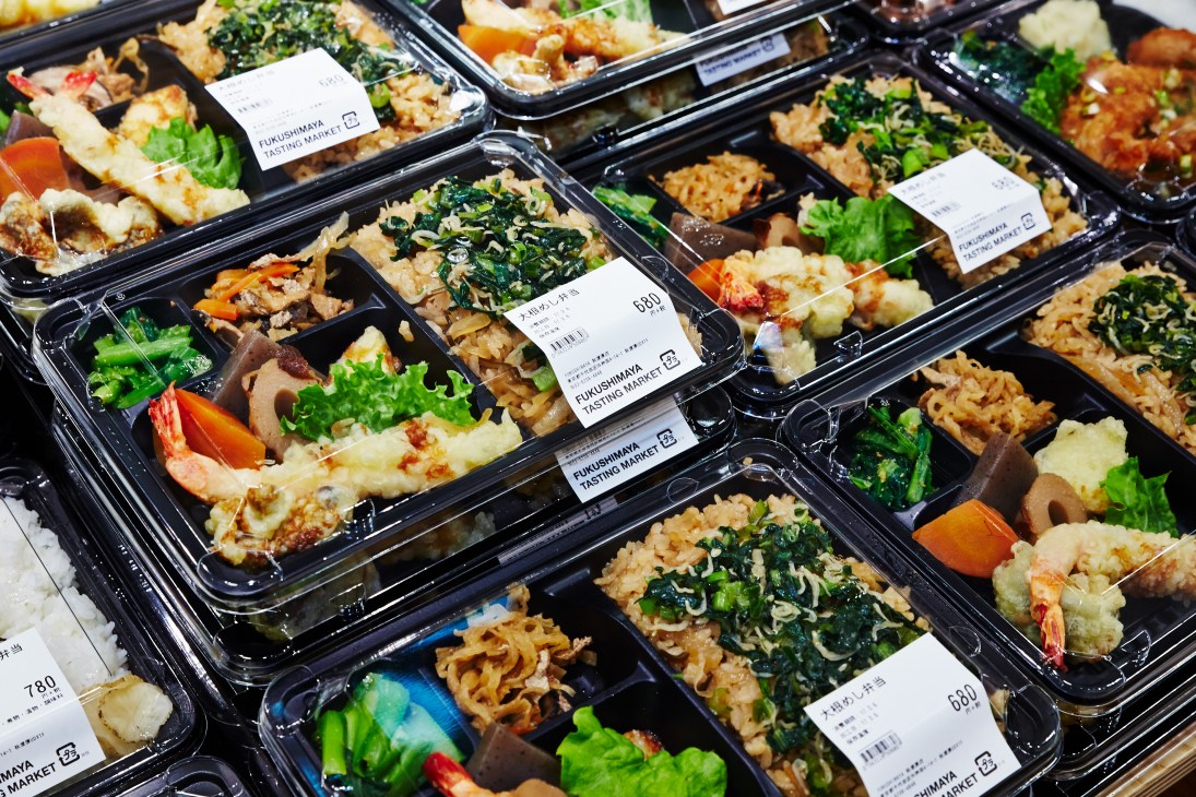 Multiple black plastic bento boxes with labeled Fukushimaya Tasting Market stickers containing prepared rice dishes with vegetables, seafood and garnishes.