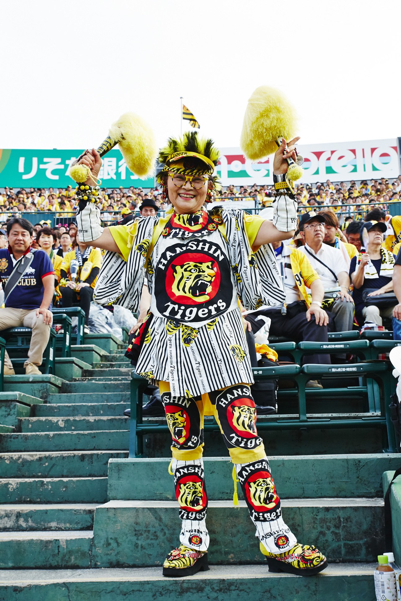 A woman in an elaborately embroidered Hanshin Tigers pinstriped uniform stands in stadium stands waving yellow pom-poms and smiling.