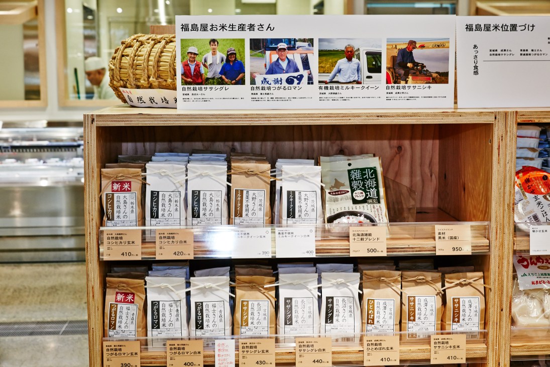 Wooden shelving display of packaged rice varieties with Japanese labels and producer profiles at Fukushimaya supermarket.