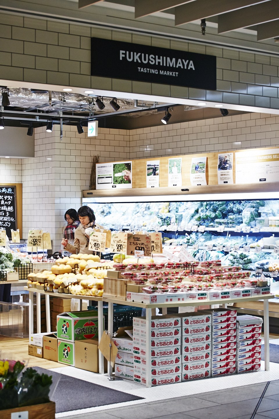 Fukushimaya Tasting Market produce counter displaying fresh fruits and vegetables with producer profile photos on the wall above.