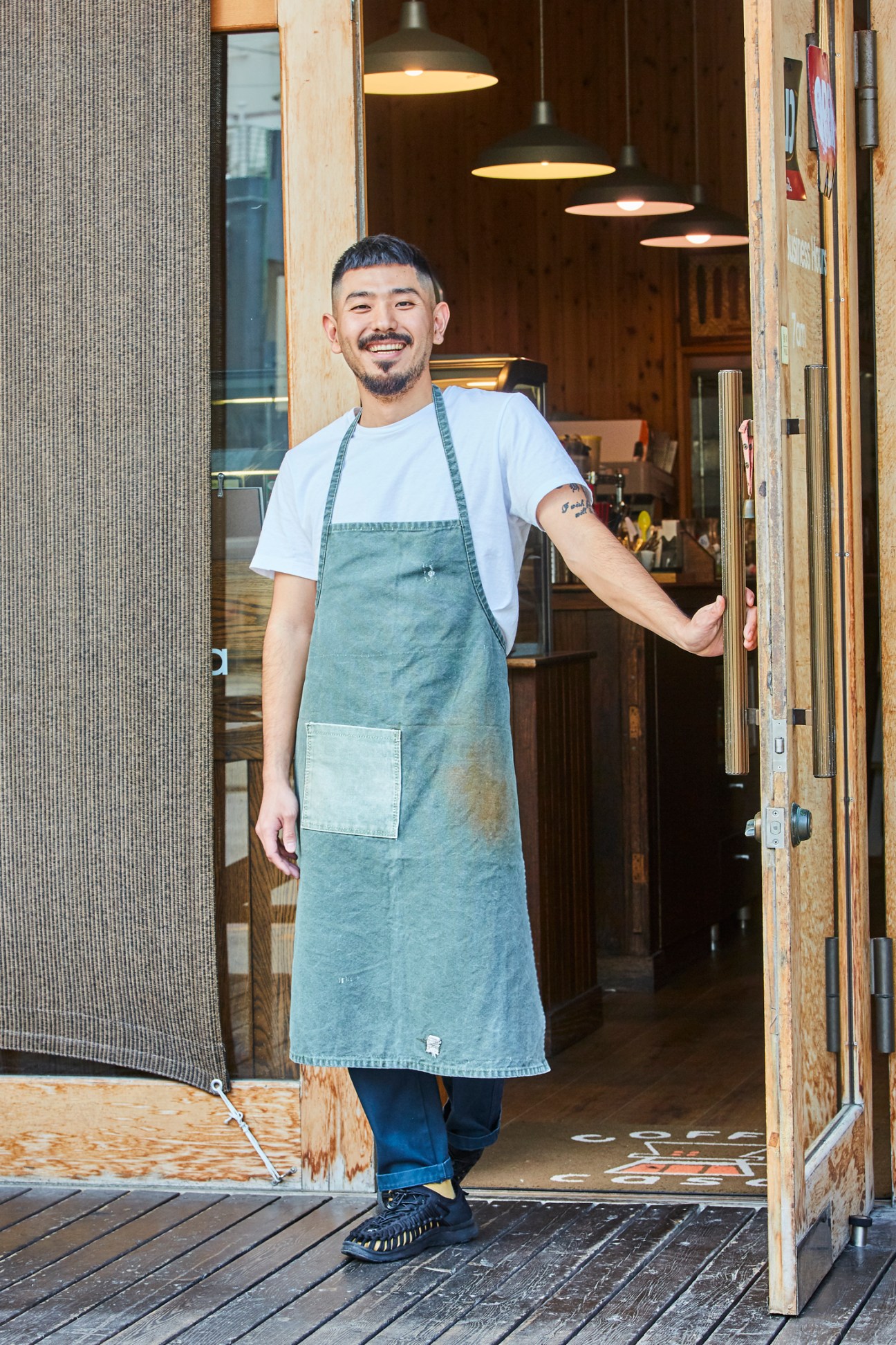 Yoshichika Hanashiro, a barista at Coffee Casa in Chatan, stands smiling in doorway wearing white t-shirt and denim apron