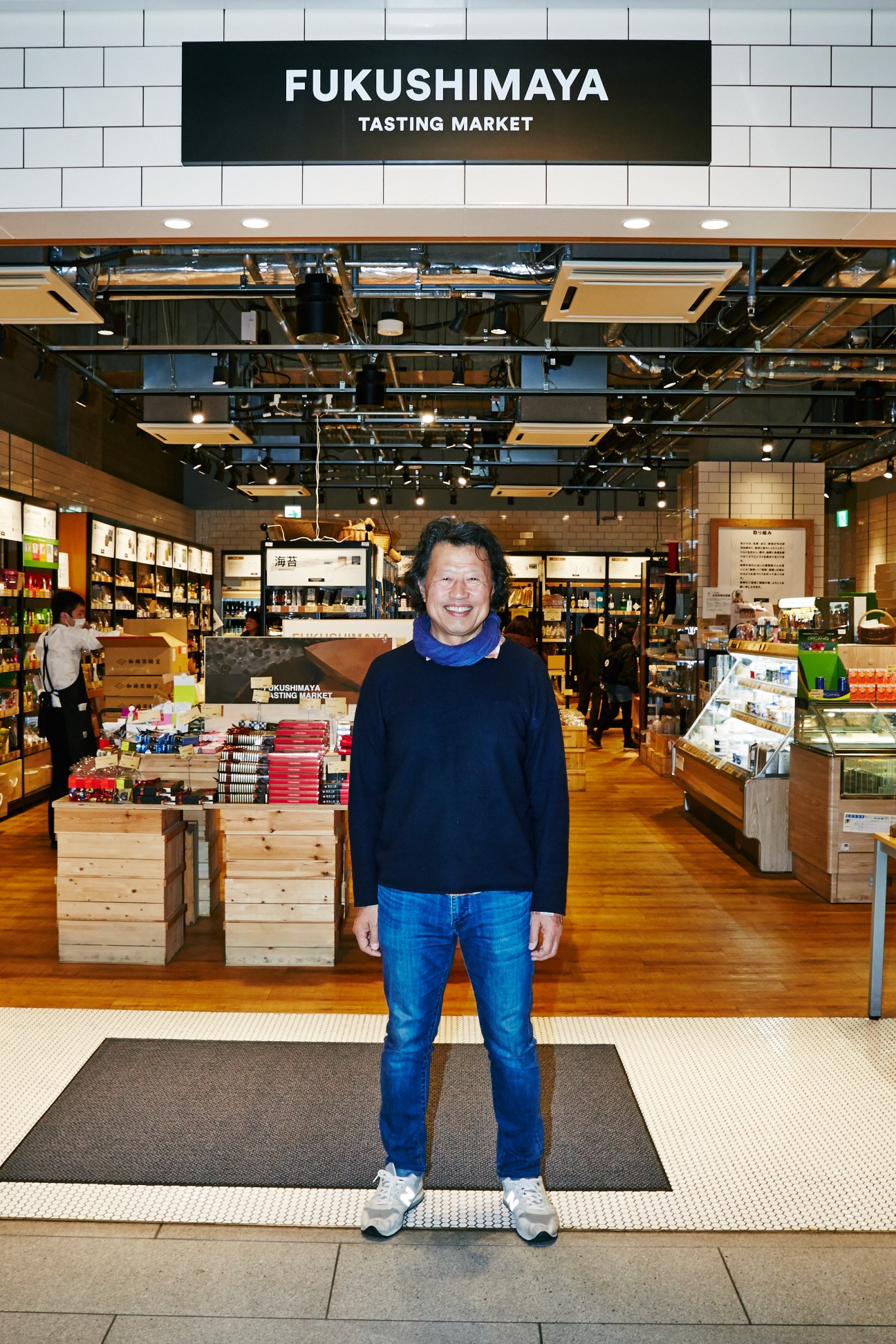 Chairman Toru Fukushima standing in Fukushimaya Tasting Market in Tokyo, a grocery store with wooden shelves and products visible behind him.