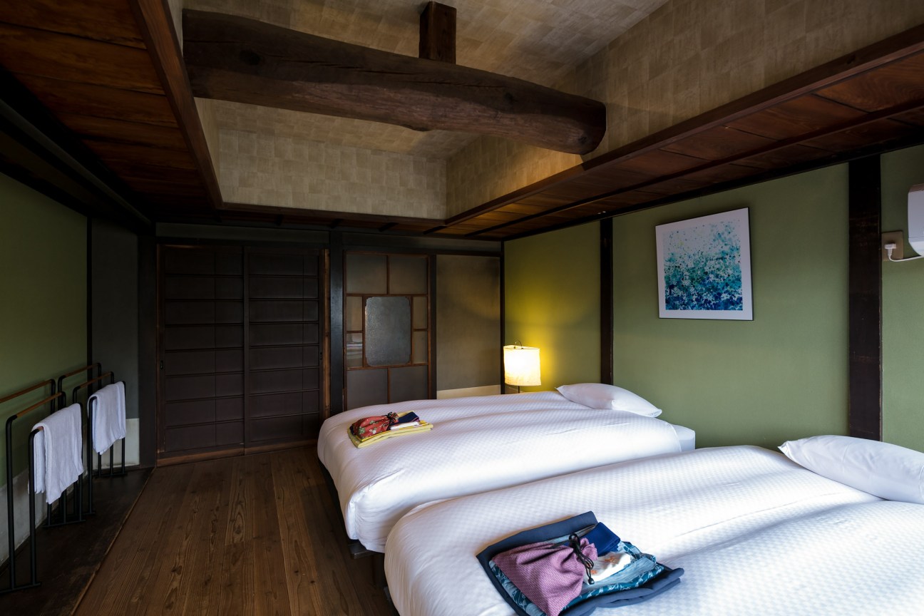 A modern Japanese bedroom with green walls, wooden beams, two beds with white linens, and traditional design elements.