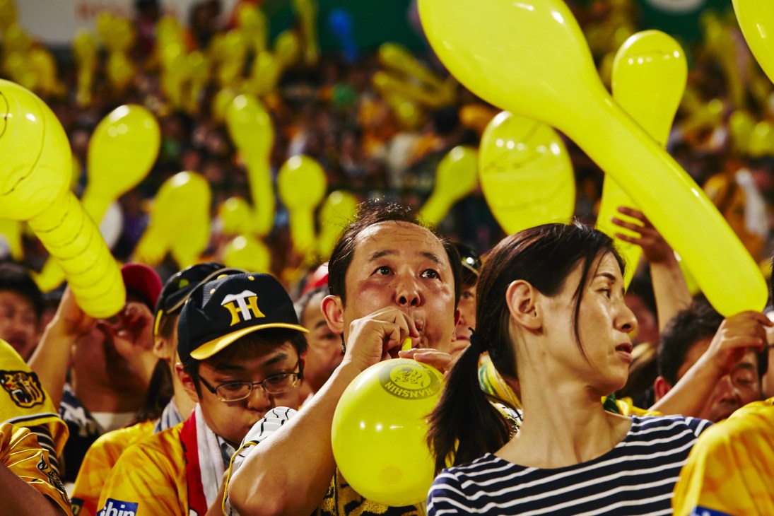 Hanshin Tigers fans in yellow uniforms holding yellow balloons and cheering in stadium stands