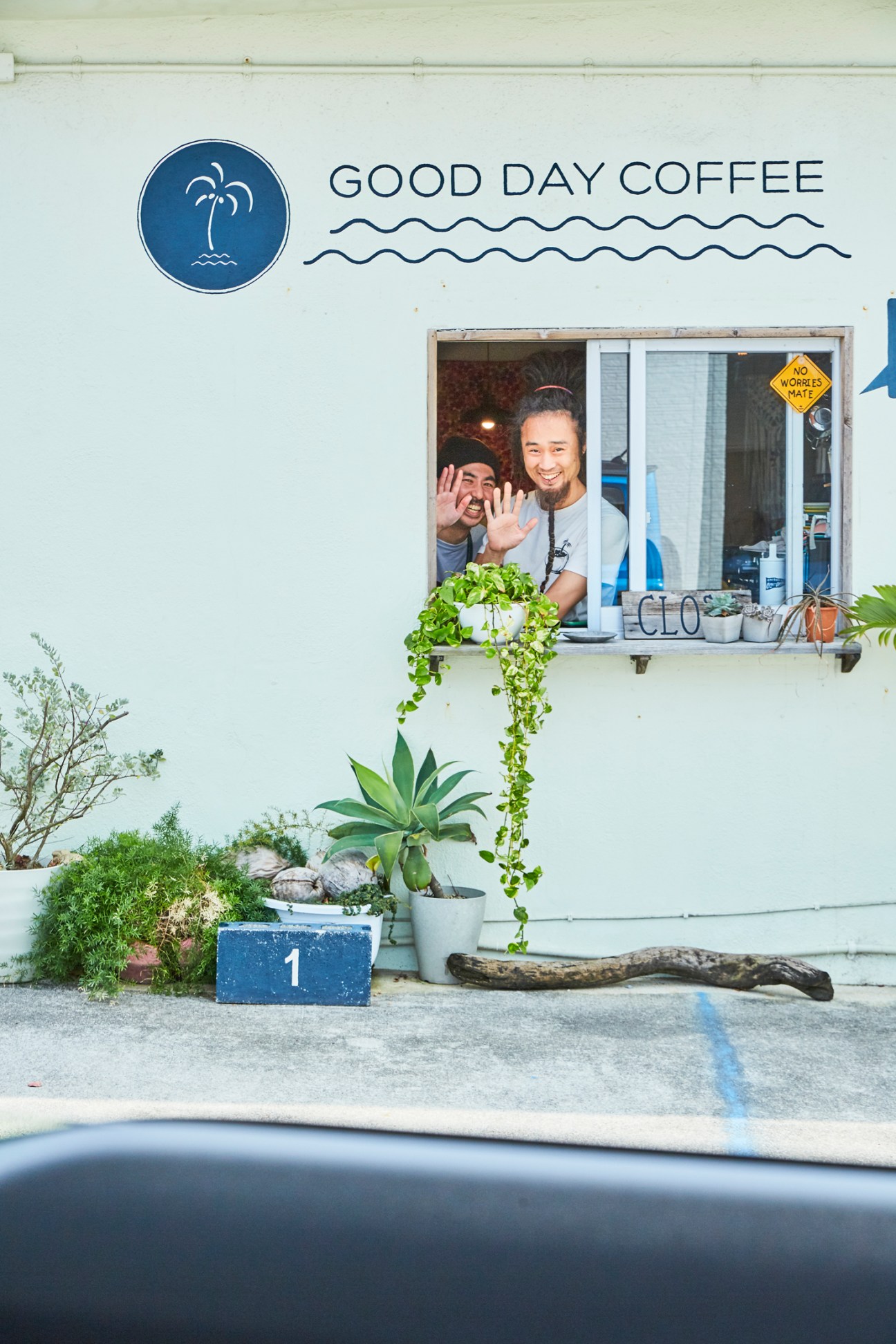 Two smiling baristas waving from service window of Good Day Coffee shop with tropical plants below